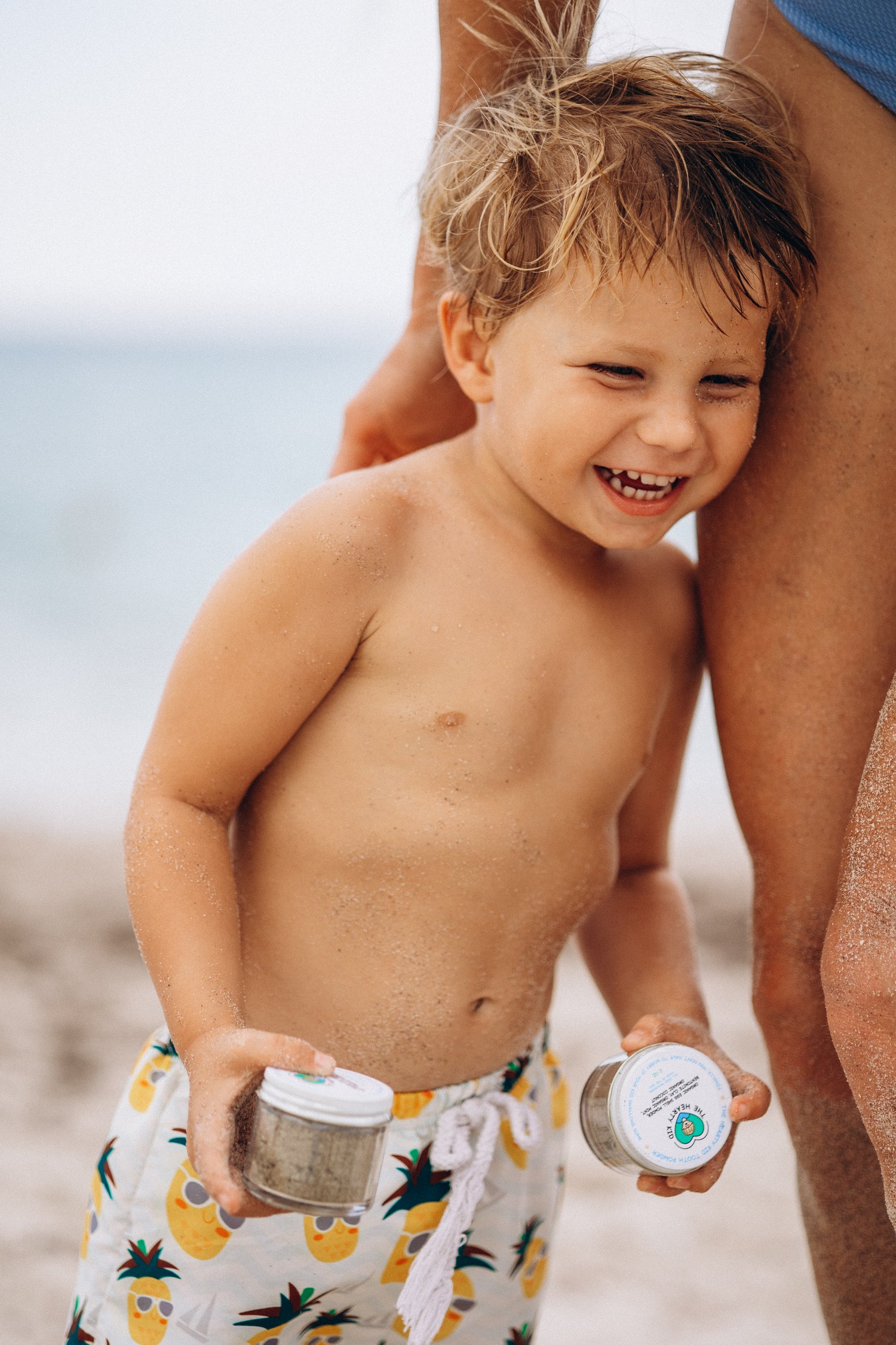 A laughing toddler at the beach holding two jars of The Hearty Kid products