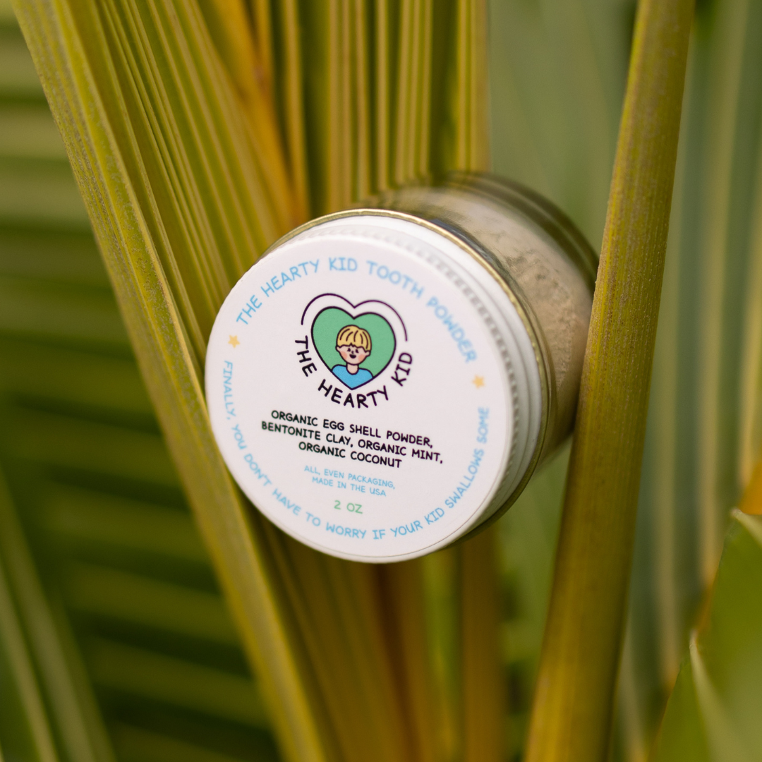 The Hearty Kid Organic Kids Tooth Powder jar resting on palm fronds